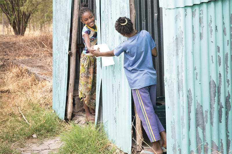 One young girl standing in the doorway of a latrine hands a roll of toilet paper to another girl in the latrine next to her. The whole structure is made of corrugated metal and wooden posts.