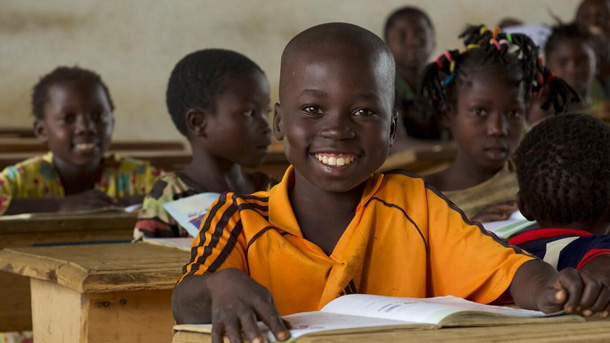 Un garçon, tout sourire, est assis à son bureau dans une salle de classe, un livre ouvert devant lui.