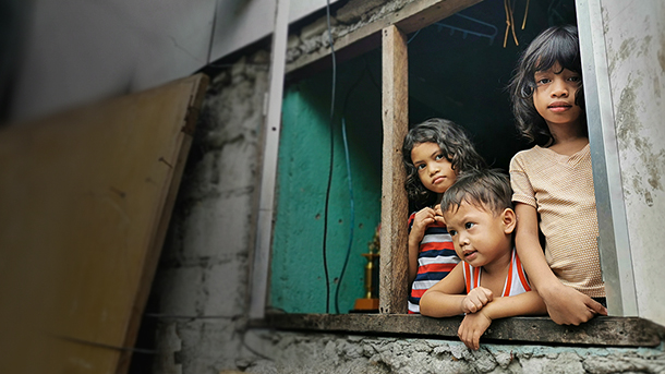 Penchés à la fenêtre d’une habitation, deux petites filles et un petit garçon regardent au-dehors.