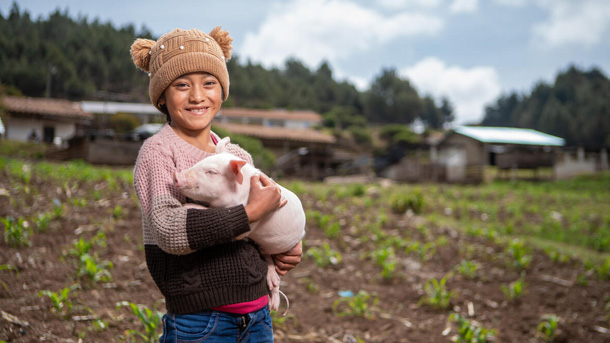 Une jeune fille souriante se tient debout dans le champ d’une ferme avec un porcelet dans les bras.