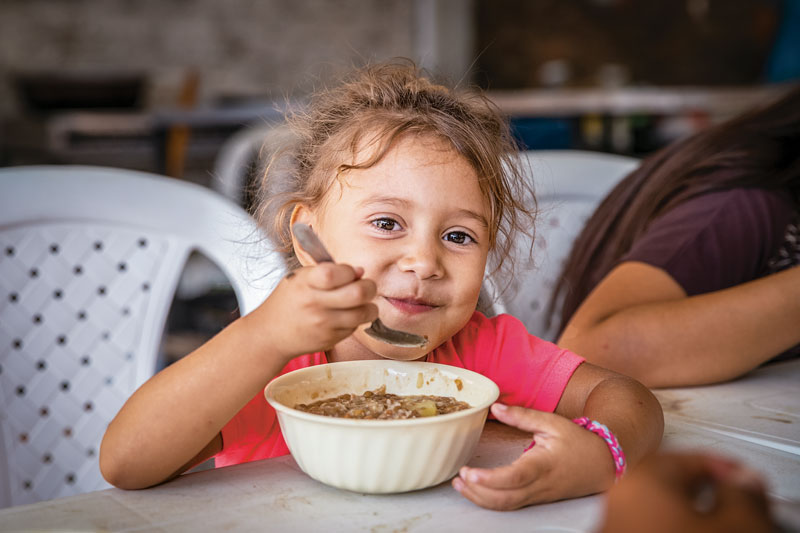 A small child is lifting a fork to their  mouth, blowing air to cool the food down before eating it.