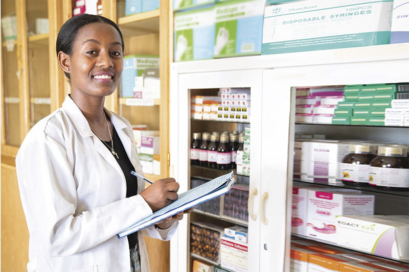 An adult woman wearing a white lab coat smiles as she stands in front of a glass-front cabinet stocked with various bottles and boxes of medicine. She holds a clipboard and a pen in each hand.