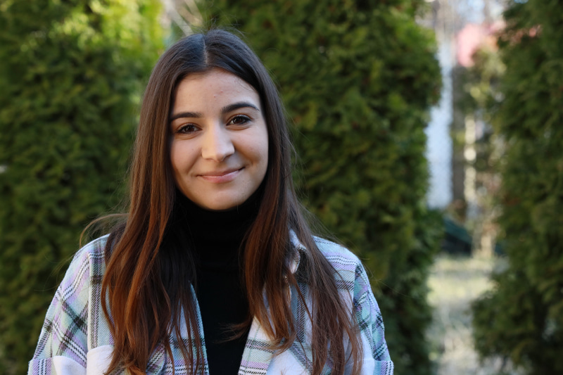 Une jeune fille aux longs cheveux bruns se tient devant de grandes haies et sourit à l’objectif.