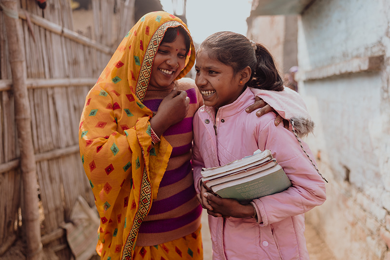 A mother in a yellow headdress wraps her arm around her daughter, who holds a stack of books close to her chest.