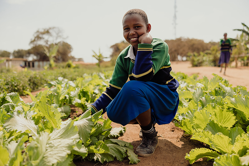 A Tanzanian girl in a green and blue school uniform kneels in a garden of lettuce, smiling at the camera.