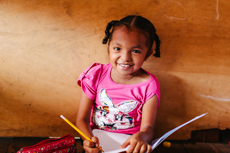 A young Peruvian girl wearing a pink t-shirt adorned with a sequined rabbit, sits and smiles as she holds a notebook and pencil.