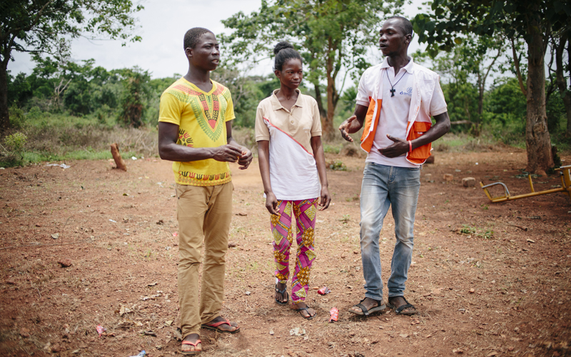 Three black young people walk in a dirt yard surrounded by greenery