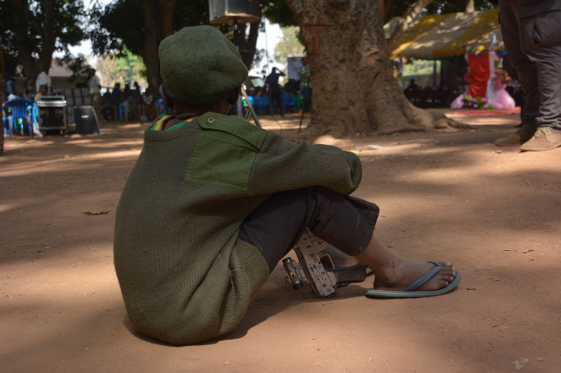 A young child soldier boy sits on a dirt ground holding a machine gun with his back to the camera.