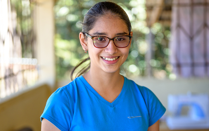 Une adolescente avec un haut bleu et des lunettes sourit en regardant l’objectif