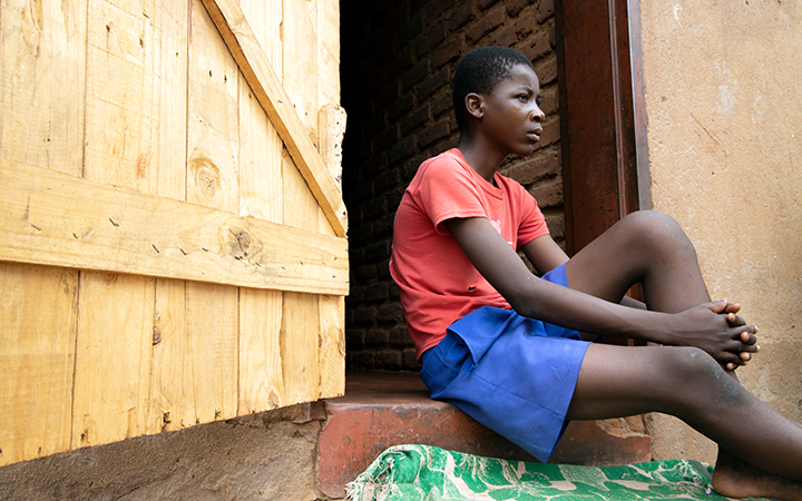Un jeune garçon en t-shirt rouge et en short bleu est assis dans l’entrée de sa maison
