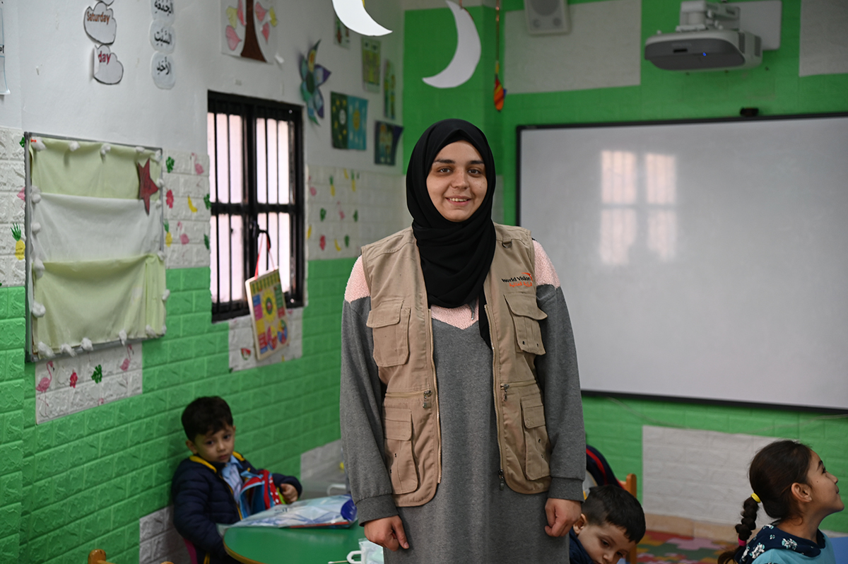 Une enseignante avec un foulard noir se tient dans une salle de classe et sourit à l’objectif. Quelques élèves sont assis à leur bureau derrière elle.