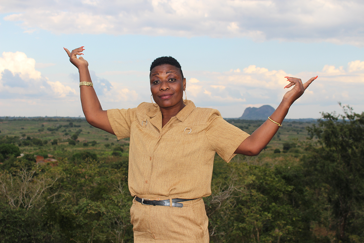 Une femme portant un uniforme beige se tient devant un paysage, les bras en l’air, dans un geste de fierté et d’accomplissement.