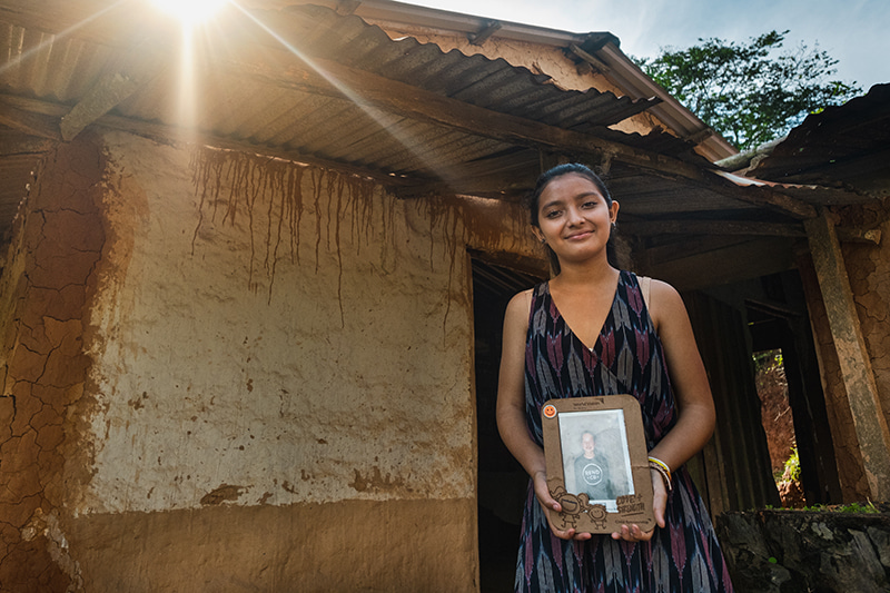 A girl stands in front of a doorway holding a framed photo of her sponsor.