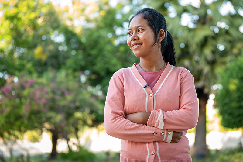 Teenage girl wearing a pink sweater stands with her arms crossed, looking to the left.