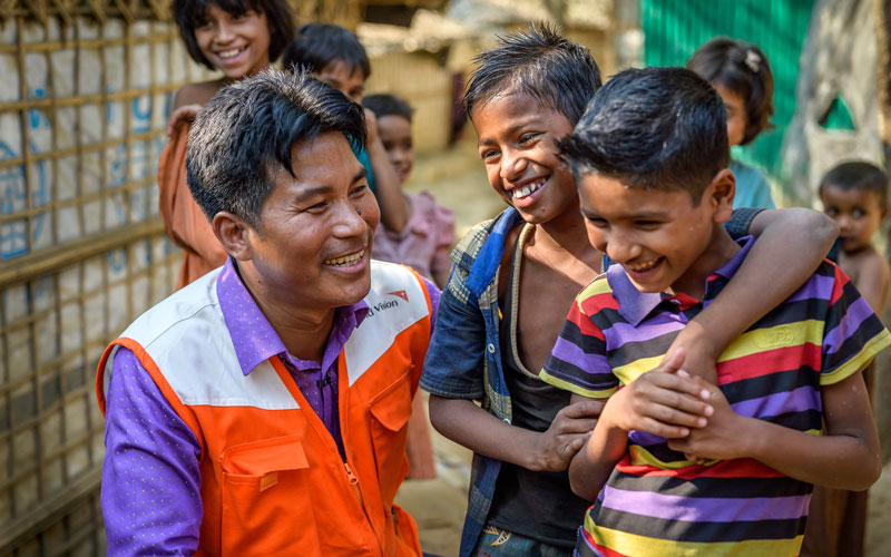 Un homme portant une veste orange et blanche Vision Mondiale sourit à deux garçons.
