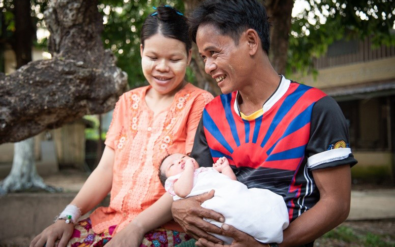 Un homme du Myanmar sourit en tenant un bébé, et une femme s’assoit à côté de lui et regarde le bébé en souriant.