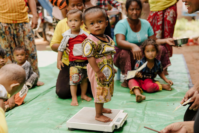 Un enfant se fait peser sur une balance. Des mères et leurs enfants sont assis autour de lui et attendent leur tour.