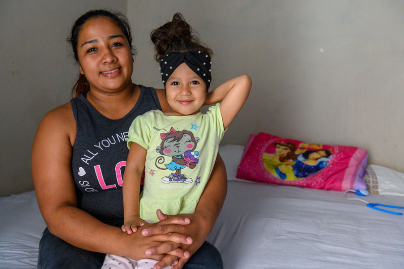 Une mère est assise sur un lit et tient dans sa fille dans ses bras tout en souriant.