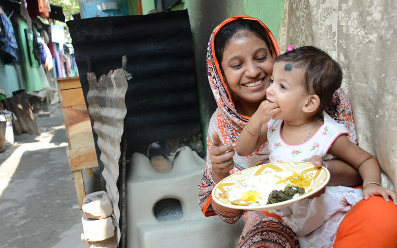A mother sits with a small girl on her lap. They are both smiling and eating from the same plate.