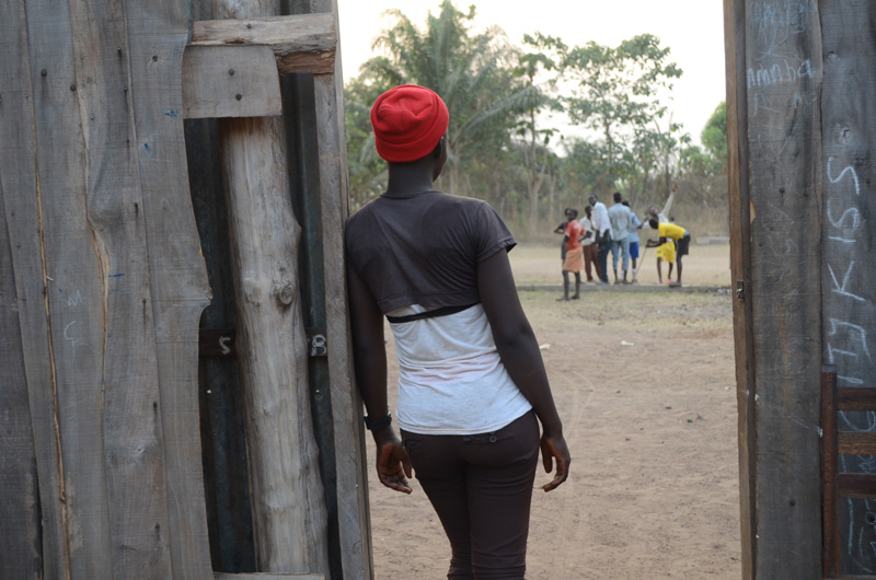 A young girl leans against a wooden door looking at a group of children playing on a dirt ground.