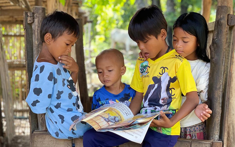 Un jeune garçon lit un livre à trois autres enfants qui l’entourent et l’écoutent.
