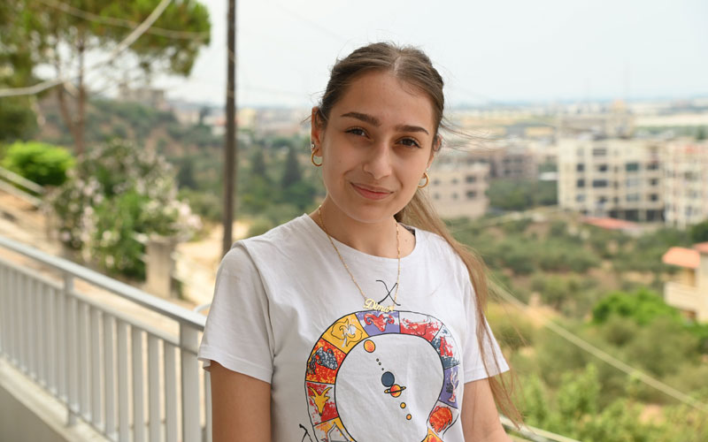 Une jeune femme portant un t-shirt blanc et un collier doré se tient sur un balcon. Elle lance à l’objectif un sourire discret.