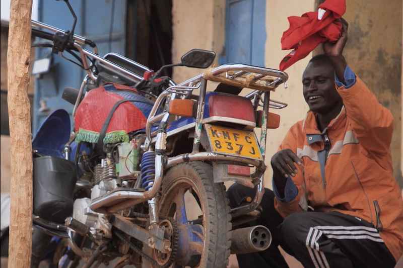 A man wearing an orange jacket, holding a red rag in the air sits beside his motorcycle.