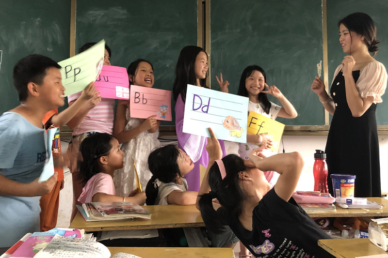 Fang, à droite, se tient face à ses élèves qui tiennent des feuilles colorées sur lesquelles est écrit l’alphabet.