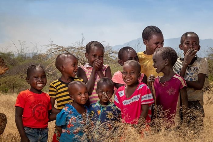 À Soin, au Kenya, dans un décor de montagnes lointaines, 10 petits enfants vêtus de différents t-shirts colorés se tiennent dans un champ, souriant devant l’appareil photo. 