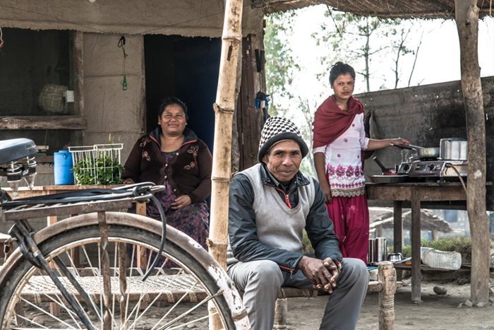 Un homme et une femme souriants sont assis devant leur modeste maison à Accham West, au Népal, avec une bicyclette au premier plan. Une deuxième femme se tient près de la femme assise et tient une mar