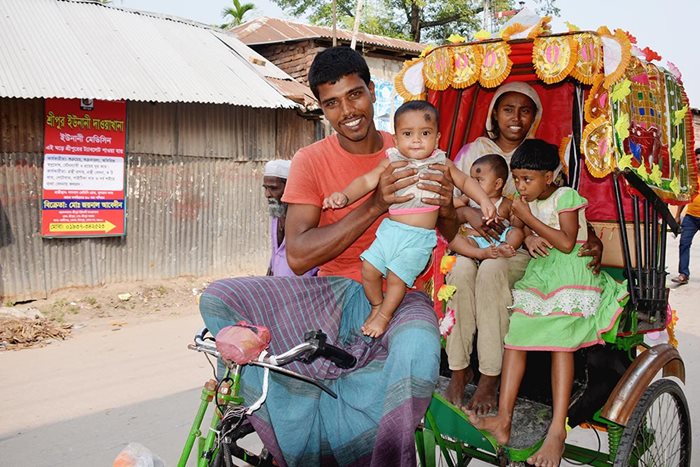 Une famille souriante de cinq personnes - un homme, une femme et trois petits enfants – à bord d’un pousse-pousse minutieusement décoré le long d'une route pavée à Bhandaria, au Bangladesh. L'homme vê