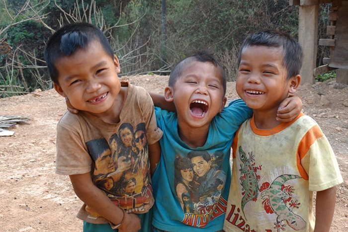 À l'extérieur d'une maison sur pilotis sur un terrain poussiéreux, trois petits garçons vêtus de t-shirts colorés se mettent les bras l’un autour de l’autre en souriant à pleines dents, dans un village au Laos.